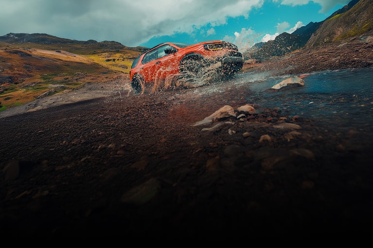 2026 Honda Passport TrailSport Elite in Sunset Orange front splashing through water on a rugged mountain trail (Mid-Size SUV).